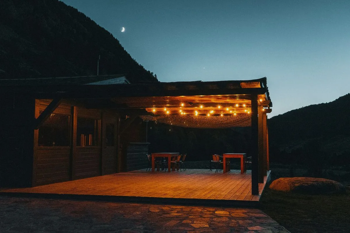 Holzterrasse mit Lichterkette in der Dämmerung, im Hintergrund ein Berghang