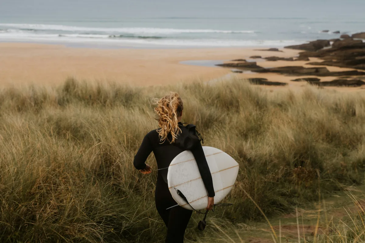A person with a surfboard walking over a dune among the grasses