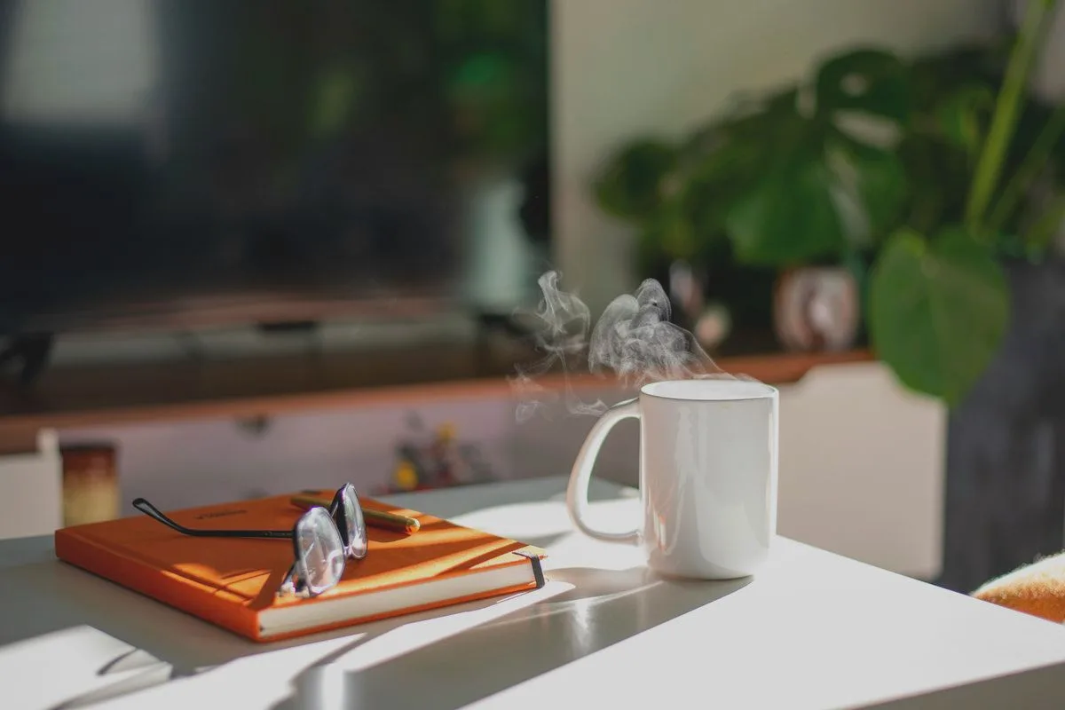A steaming mug and a notebook with glasses on a table in the morning sun